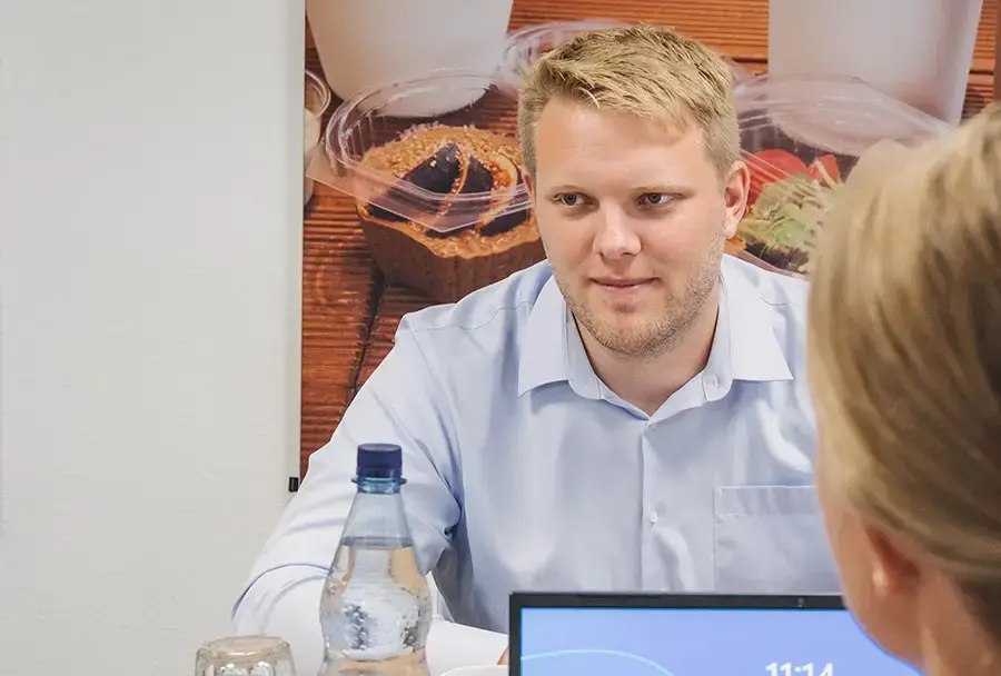 Ein Mann mit blauer Hemd sitzt an einem Tisch während eines Meetings mit einer Wasserflasche und einem Laptop.