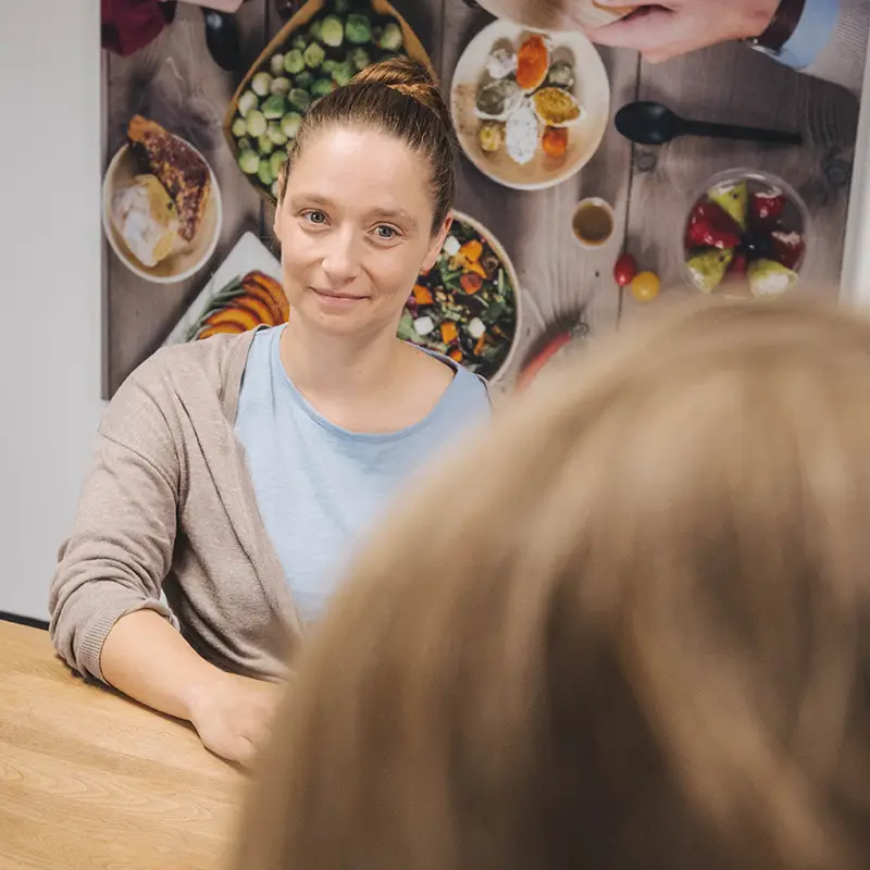 Frau mit braunen Haaren sitzt an einem Tisch und spricht mit einer anderen Person.