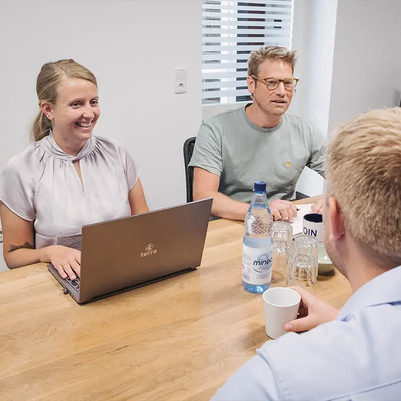 Drei Personen in einem Meeting, eine Person arbeitet am Laptop, zwei weitere unterhalten sich am Tisch.